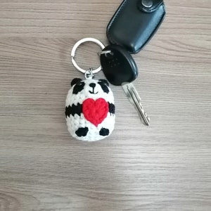 May include: A crocheted panda keychain holding a red heart. The keychain is attached to a silver key ring and a black car key.