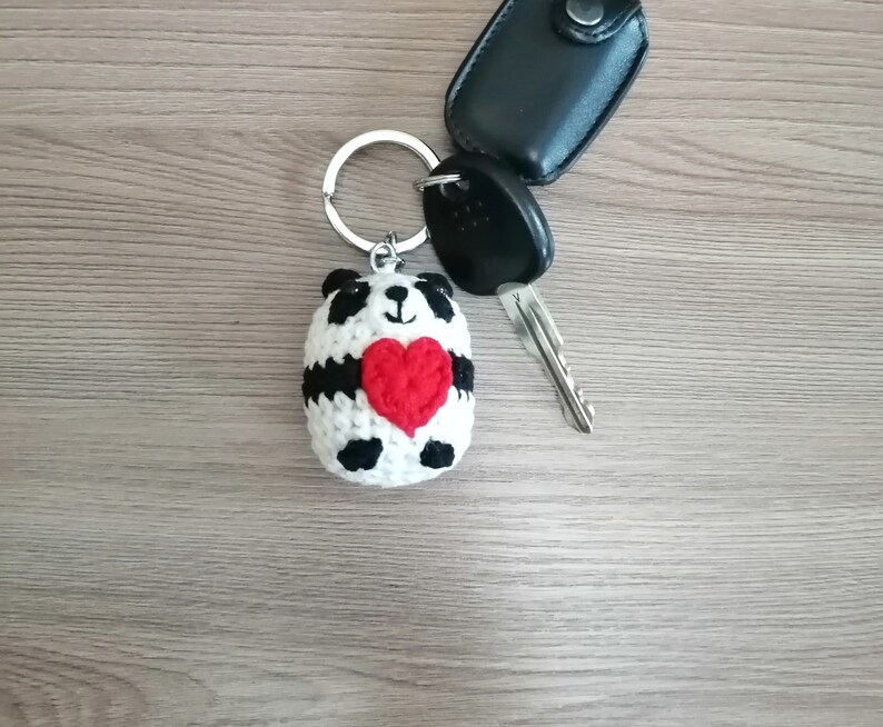 May include: A crocheted panda keychain holding a red heart. The keychain is attached to a silver key ring and a black car key.