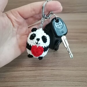 May include: A crocheted panda bear keychain holding a red heart. The keychain is attached to a silver key ring.