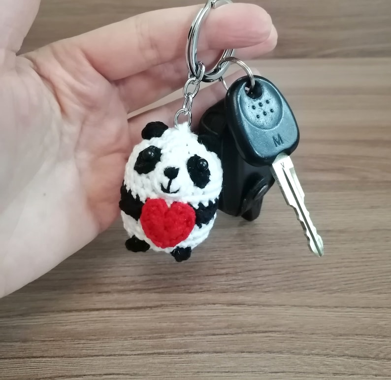 May include: A crocheted panda bear keychain holding a red heart. The keychain is attached to a silver key ring.