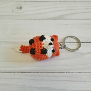 Puede incluir: Un llavero de panda rojo tejido a crochet con ojos negros y una cara blanca. El llavero está unido a un llavero plateado.