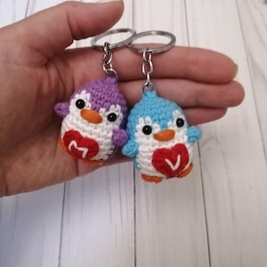 May include: Two crocheted penguin keychains with red hearts. The penguin on the left is purple and the penguin on the right is blue. Each heart has a letter on it, "M" and "V".