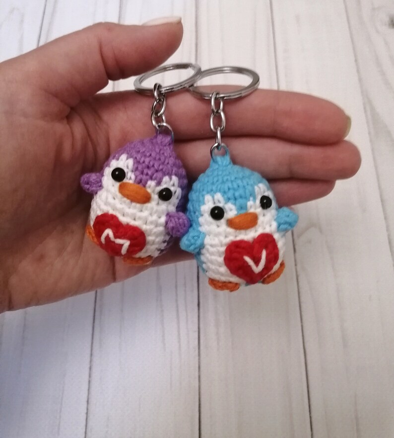 May include: Two crocheted penguin keychains with red hearts. The penguin on the left is purple and the penguin on the right is blue. Each heart has a letter on it, "M" and "V".