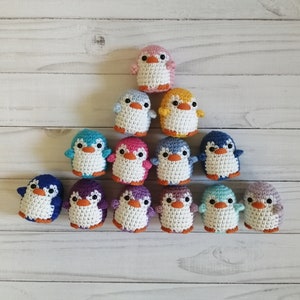 May include: A collection of 13 crocheted penguin ornaments in various colors, including pink, blue, yellow, purple, and green. The penguins are arranged in a pyramid shape on a white wooden surface.