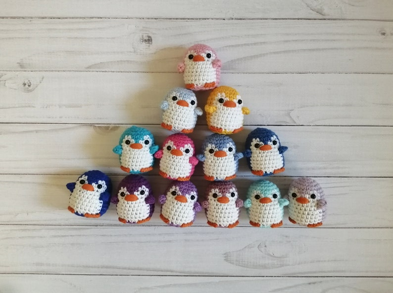 May include: A collection of 13 crocheted penguin ornaments in various colors, including pink, blue, yellow, purple, and green. The penguins are arranged in a pyramid shape on a white wooden surface.