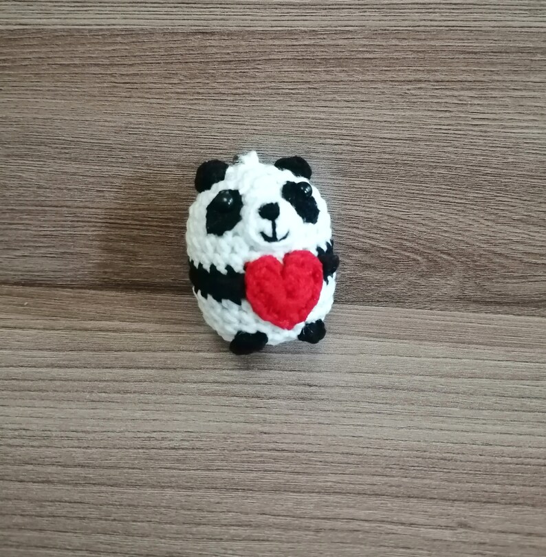 May include: A crocheted panda holding a red heart. The panda is white with black ears, eyes, and paws.
