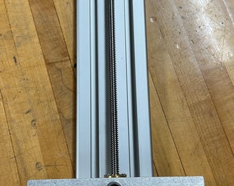 May include: A silver metal linear actuator with a threaded rod and a knob at the bottom. The actuator has two silver metal plates on the top and bottom. The top plate has the text "NYLE NEW YORK LASER ENGRAVING" on it. The bottom plate has the text "NYLE NEW YORK LASER ENGRAVING" on it.