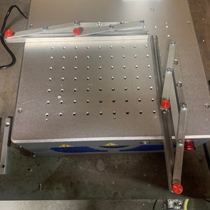 May include: A silver metal work surface with a grid of holes. Two silver metal arms with red knobs are attached to the surface. The arms are in a raised position.
