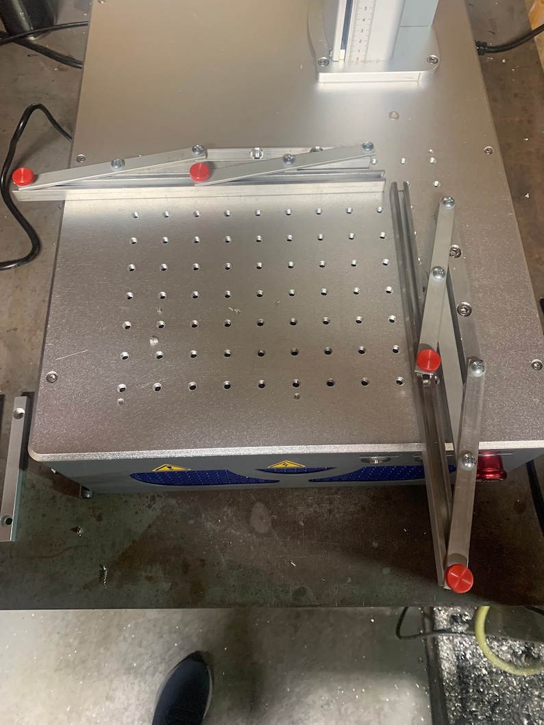 May include: A silver metal work surface with a grid of holes. Two silver metal arms with red knobs are attached to the surface. The arms are in a raised position.