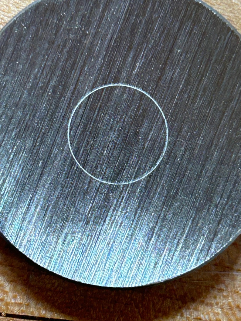 May include: A close-up of a round, silver metal disc with a white circle etched in the center.