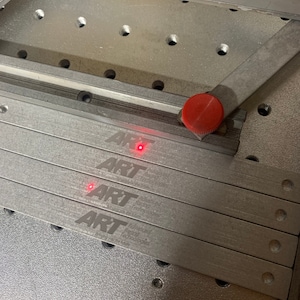 May include: Four silver metal strips with the text "ART" and "Advanced Rail Technologies" engraved on them. The strips are stacked on top of each other and are sitting on a grey metal surface.