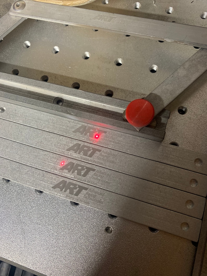 May include: Four silver metal strips with the text "ART" and "Advanced Rail Technologies" engraved on them. The strips are stacked on top of each other and are sitting on a grey metal surface.