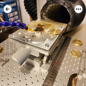 May include: A close-up of a silver and black machine with a round knob and a ruler marked in inches. The machine has a circular platform with a small, round, brown object on it. The machine is on a gray, textured surface with a grid pattern.