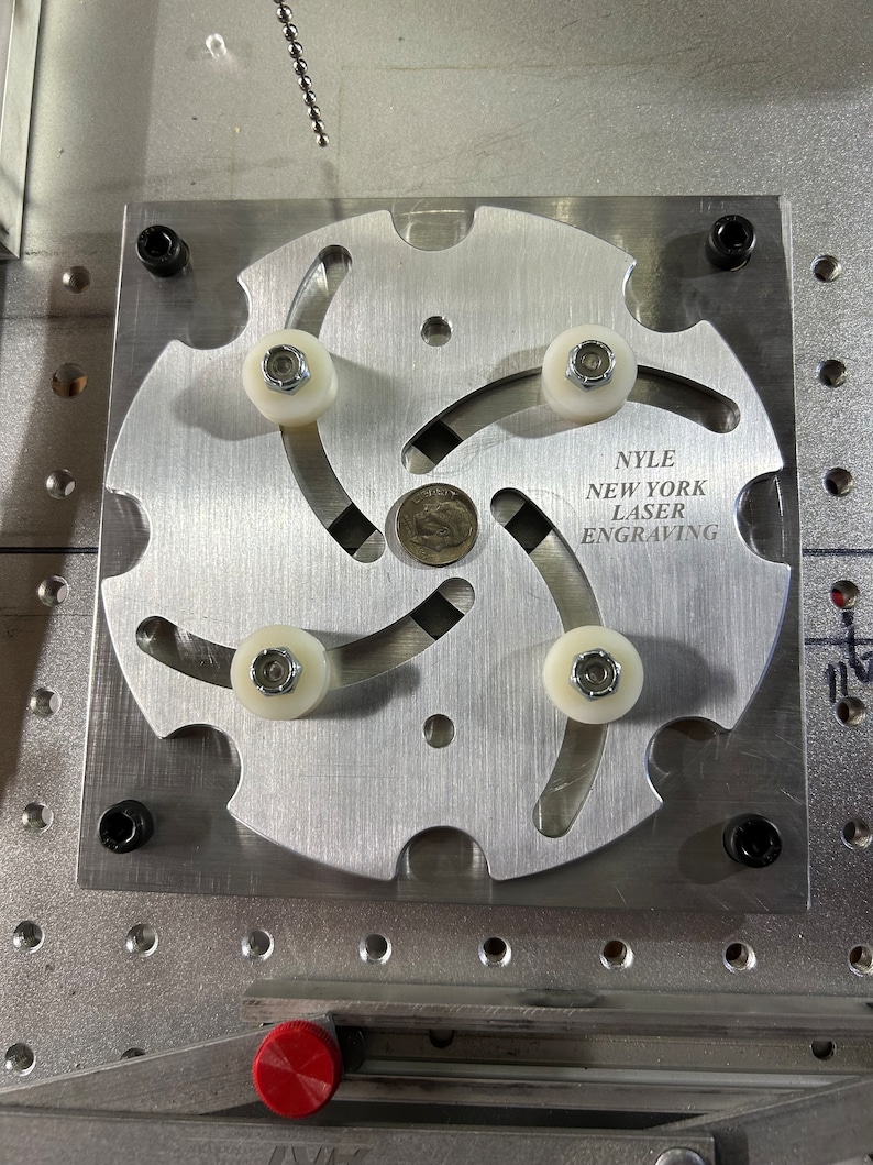 May include: A silver metal rotary indexing table with four white plastic rollers and the text "NYLE NEW YORK LASER ENGRAVING" on the face.