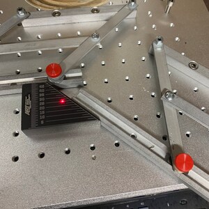 May include: A silver metal work surface with a grid pattern of holes. Two silver metal arms with red knobs are attached to the surface. A black protractor with a red laser pointer is positioned on the surface.