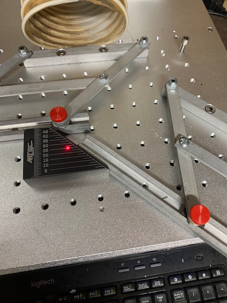 May include: A silver metal work surface with a grid pattern of holes. Two silver metal arms with red knobs are attached to the surface. A black protractor with a red laser pointer is positioned on the surface.