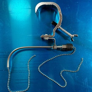 May include: A silver metal clamp with a chain and a curved arm. The clamp is designed to attach to a surface and the arm can be adjusted to hold an object.