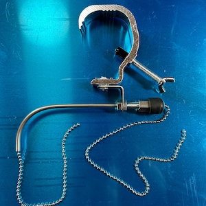 May include: A silver metal clamp with a chain and a curved arm. The clamp is designed to attach to a surface and the arm can be adjusted to hold an object.
