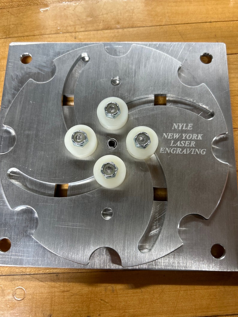 May include: A silver metal plate with a circular design featuring five white plastic rollers with silver metal screws. The plate has the text "NYLE NEW YORK LASER ENGRAVING" engraved on it.