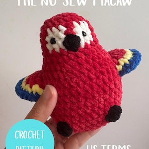 Puede incluir: Un guacamayo rojo tejido a mano con detalles blancos, negros, amarillos y azules. El texto "THE NO-SEW MACAW" está en la parte superior, y un círculo azul con "CROCHET PATTERN" y "US TERMS" está en la parte inferior. El pájaro está sostenido en una mano.