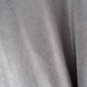 Puede incluir: Primer plano de una tela gris texturizada con un sutil patrón similar a escamas. El material está drapeado, creando pliegues y sombras que resaltan la textura y el brillo de la tela. La apariencia general es suave y elegante.