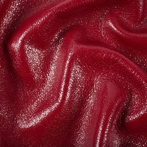 May include: Close-up of a textured, glossy, deep red leather material. The surface has a slight sheen and visible grain, with folds and curves creating shadows and highlights. The overall appearance suggests a high-quality material.