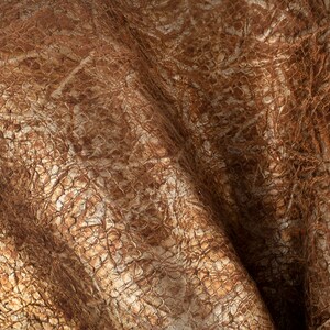 May include: Close-up of a brown leather fabric with a textured, embossed pattern. The fabric has a subtle shimmer and a wrinkled, crinkled appearance.