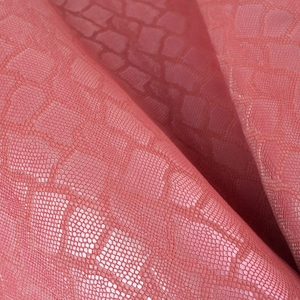 Op de afbeelding: Close-up van een gevouwen stuk roze imitatieleer met een reptielachtig reliëfpatroon. Het materiaal heeft een getextureerd oppervlak en een subtiele glans, met highlights en schaduwen.