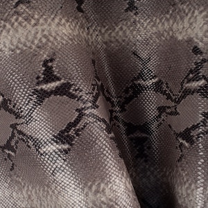 Puede incluir: Primer plano de una tela de piel de serpiente gris y negra con un patrón texturizado. La tela está doblada y muestra los intrincados detalles de las escamas.