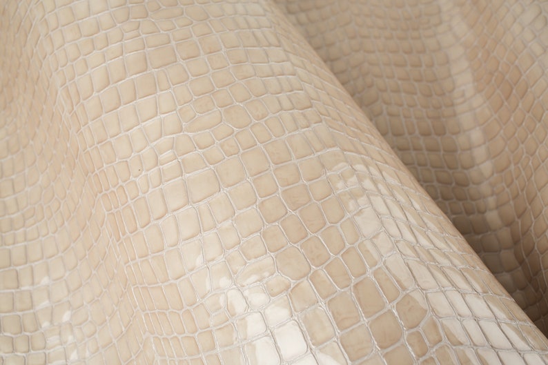 Puede incluir: Primer plano de una tela de cuero beige con relieve de cocodrilo. La textura es detallada y realista.