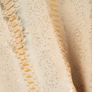 Puede incluir: Primer plano de una tela color crema con un diseño de piel de serpiente. El diseño presenta escamas doradas y plateadas, creando un efecto texturizado y brillante. La tela está ligeramente arrugada, añadiendo profundidad.