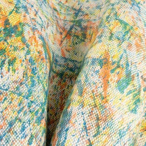 Op de afbeelding: Close-up van een textiel met een kleurrijk abstract ontwerp. De stof heeft een rasterpatroon dat is overlegd met spetters blauwe, oranje en gele verf, waardoor een levendige en artistieke uitstraling ontstaat. De stof is gevouwen en toont diepte en textuur.