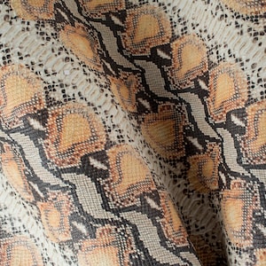 Op de afbeelding: Close-up van een stof met een slangenhuidpatroon. De stof is lichtbruin van kleur met zwarte en oranje accenten.