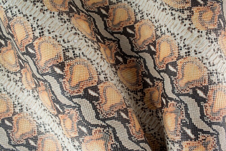 Op de afbeelding: Close-up van een stof met een slangenhuidpatroon. De stof is lichtbruin van kleur met zwarte en oranje accenten.