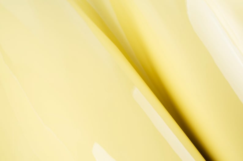 May include: A close-up of a smooth, pale yellow fabric with a subtle sheen. The fabric is folded in a diagonal pattern, creating a soft, abstract design.