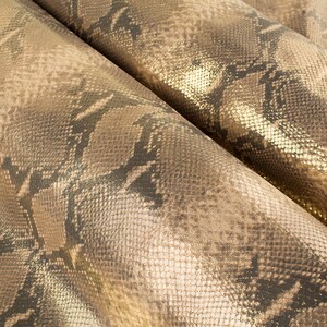 May include: Close-up of a gold and brown snakeskin fabric with a metallic sheen. The fabric has a textured surface and a repeating pattern of scales.