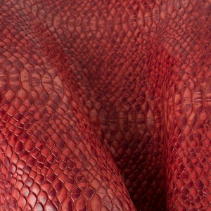 May include: Red snakeskin textured fabric with a subtle sheen. The fabric is folded and draped, showing the texture and pattern.