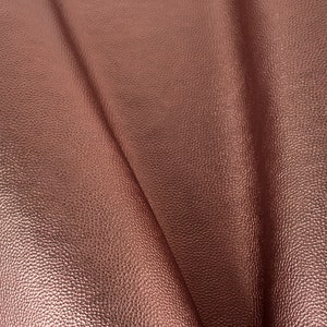 May include: Close-up of a sheet of textured, rose gold-colored faux leather. The material is folded, showing the grain and subtle variations in the metallic finish. The surface has a dimpled appearance.