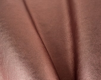 May include: Close-up of a sheet of textured, rose gold-colored faux leather. The material is folded, showing the grain and subtle variations in the metallic finish. The surface has a dimpled appearance.