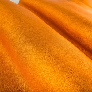 May include: Close-up of a textured, orange leather material. The leather is folded and has a soft, smooth surface.