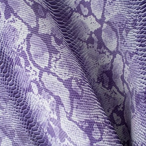 Puede incluir: Primer plano de una tela texturizada con un patrón de piel de serpiente. La tela presenta un diseño repetitivo de escamas moradas y blancas, creando un efecto realista de piel de reptil. El material está drapeado, mostrando pliegues y sombras.