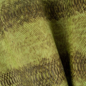 以下が含まれることがあります： 緑色のヘビ革生地で、テクスチャのあるパターンが施されています。生地は半分に折り畳まれており、両側にパターンが表示されています。