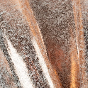 Puede incluir: Primer plano de una tela metálica de color oro rosa con una superficie texturizada y agrietada. La tela tiene un brillo sutil y una apariencia ligeramente arrugada.