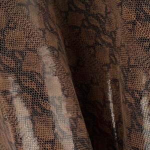 Op de afbeelding: Close-up van een textiel met een slangenpatroon. Het materiaal heeft een mix van bruine, beige en zwarte tinten, waardoor een realistisch reptielhuideffect ontstaat. De stof is gevouwen en toont de textuur en glans.