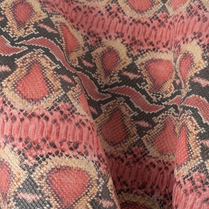 May include: Close-up of a pink, black and beige snakeskin pattern fabric. The fabric has a textured surface and a repeating pattern.