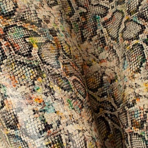 May include: Close-up of a beige fabric with a snakeskin pattern. The fabric has a textured surface and is decorated with colorful paint splatters.