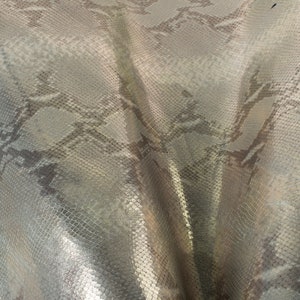 May include: Close-up of a silver snakeskin fabric with a textured pattern. The fabric has a subtle sheen and a slightly wrinkled appearance.