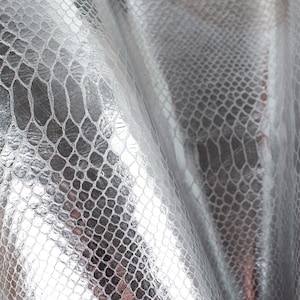 May include: Silver faux snakeskin fabric with a textured, scaly pattern. The fabric is folded and draped, showing the texture and sheen of the material.