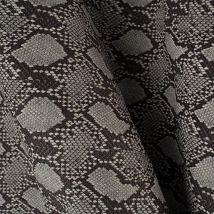 May include: Close-up of a fabric with a snakeskin pattern. The material features a repeating design of grey and black scales, creating a textured appearance. The material is draped, showing folds and shadows.