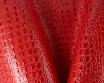 May include: Close-up of red alligator-textured leather. The material has a glossy finish and a repeating pattern of rectangular scales. The leather is folded, creating shadows and highlighting the texture. This material is suitable for various crafting projects.